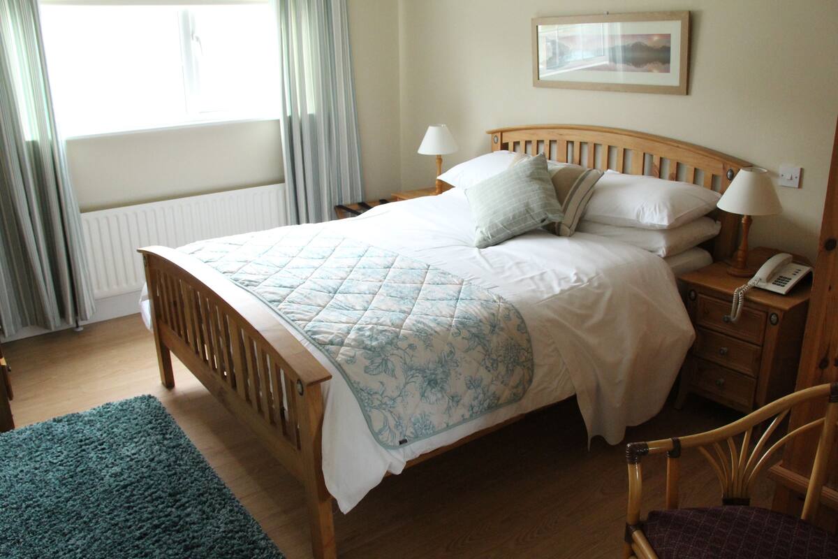 A neatly made bed with a floral duvet, a bedside table with a lamp, and a chair in a well-lit room with a window and curtains.