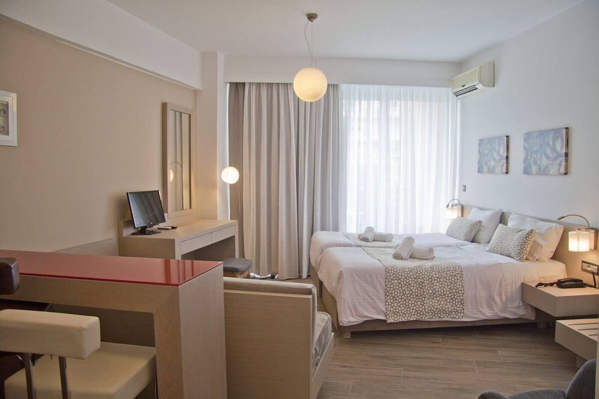 A hotel room with a desk, two beds, a chair, a desk lamp, and artwork on the wall.