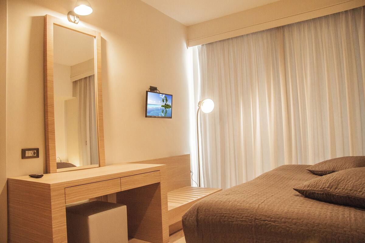 A hotel room with a wooden desk, a large mirror, a television mounted on the wall, and a bed with brown bedding.