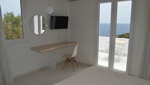 Une chambre avec un grand lit, un téléviseur fixé au mur, une table de chevet en bois, une chaise et une vue sur la mer par la fenêtre.