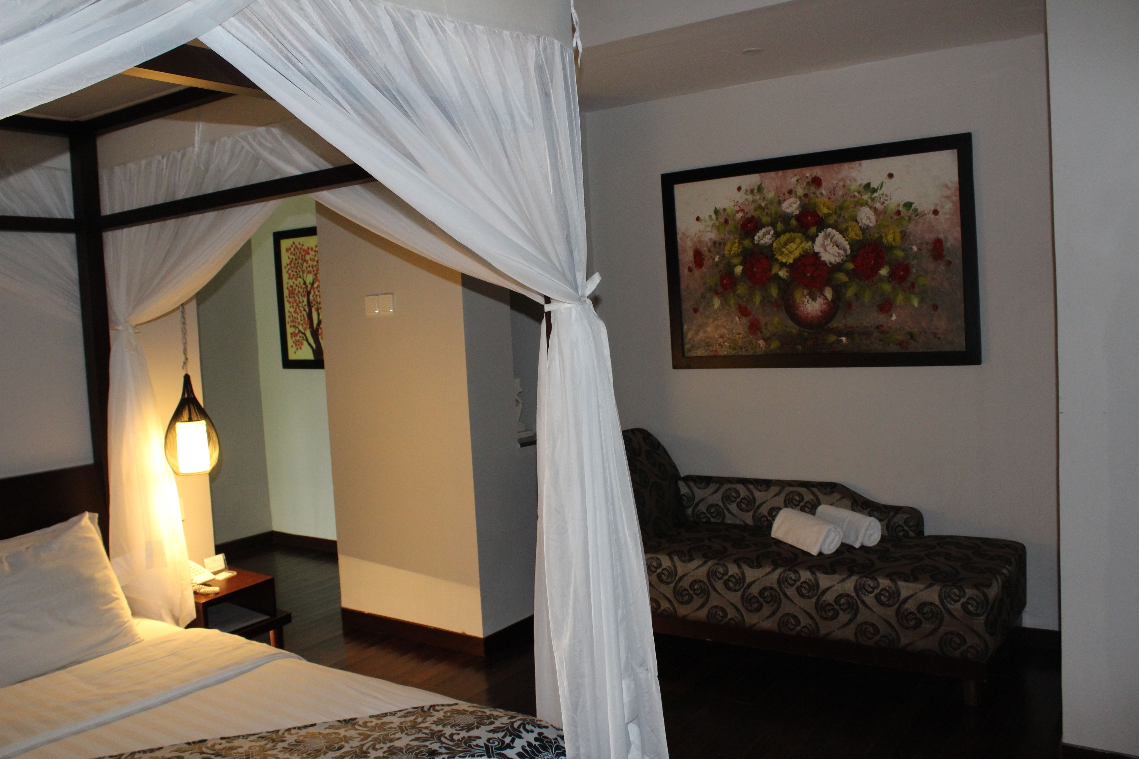 A bedroom with a four-poster bed, a sofa, a painting, and a nightstand with a lamp.