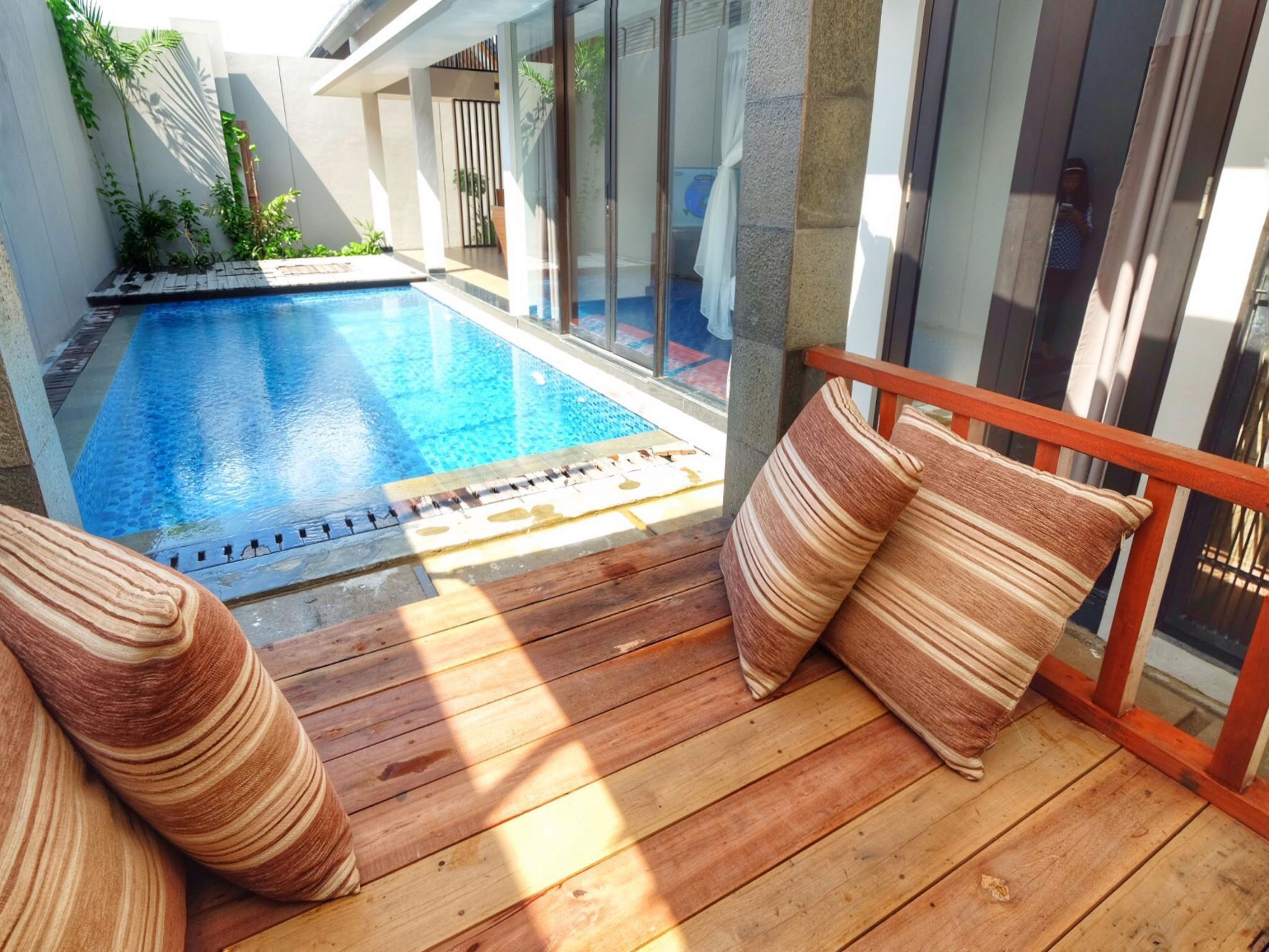 A wooden deck with a pool, cushioned seating, and a glass balustrade.
