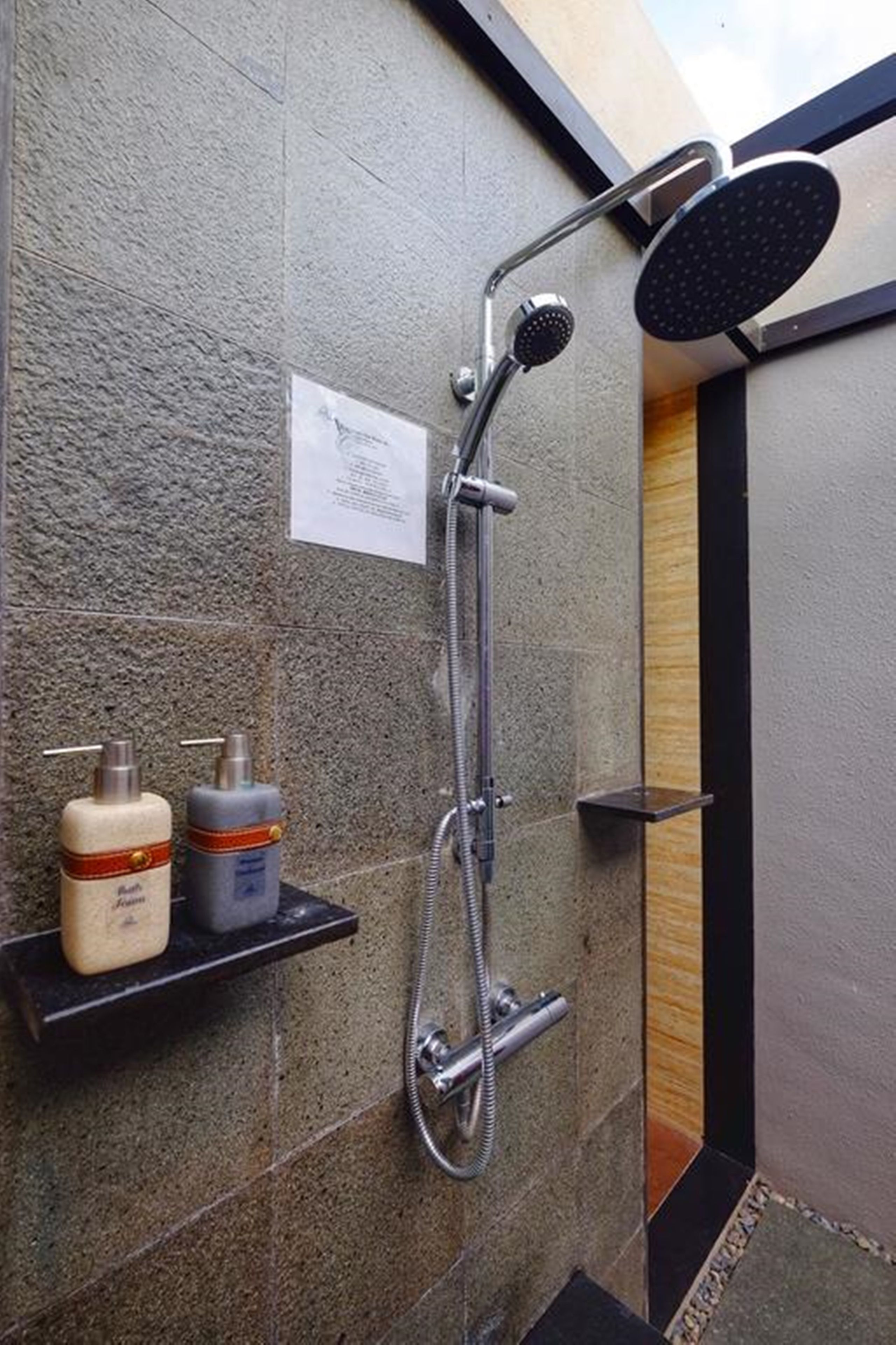A shower with a handheld showerhead and a wall-mounted head. There are two bottles on a shelf, one labeled "Kleenex" and the other "Lotion."