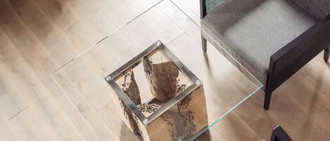 A modern room with a glass coffee table featuring a wooden centerpiece, a grey sofa, and a wooden floor.