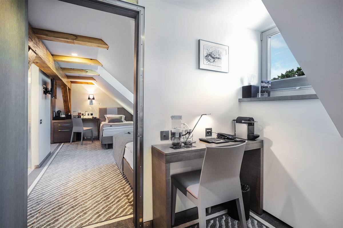 A modern hotel room with a desk, chair, and a bed. There is a window with a view of the sky and trees.
