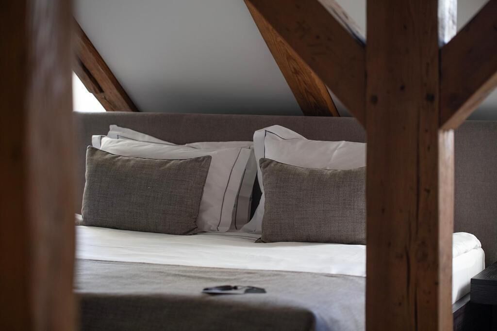 A bedroom with a sloped ceiling, wooden beams, a bed with grey bedding, and a pillow.