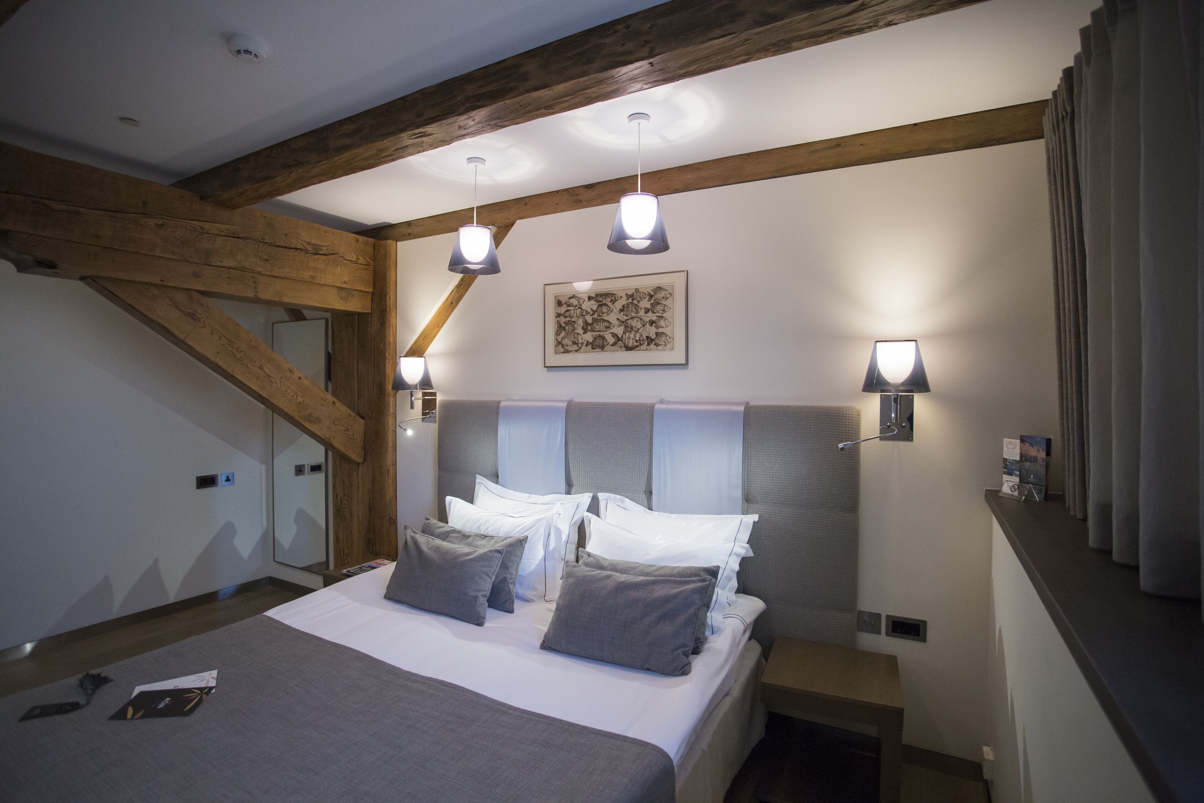 A modern hotel room with a large bed, wooden beams, and wall-mounted lighting.