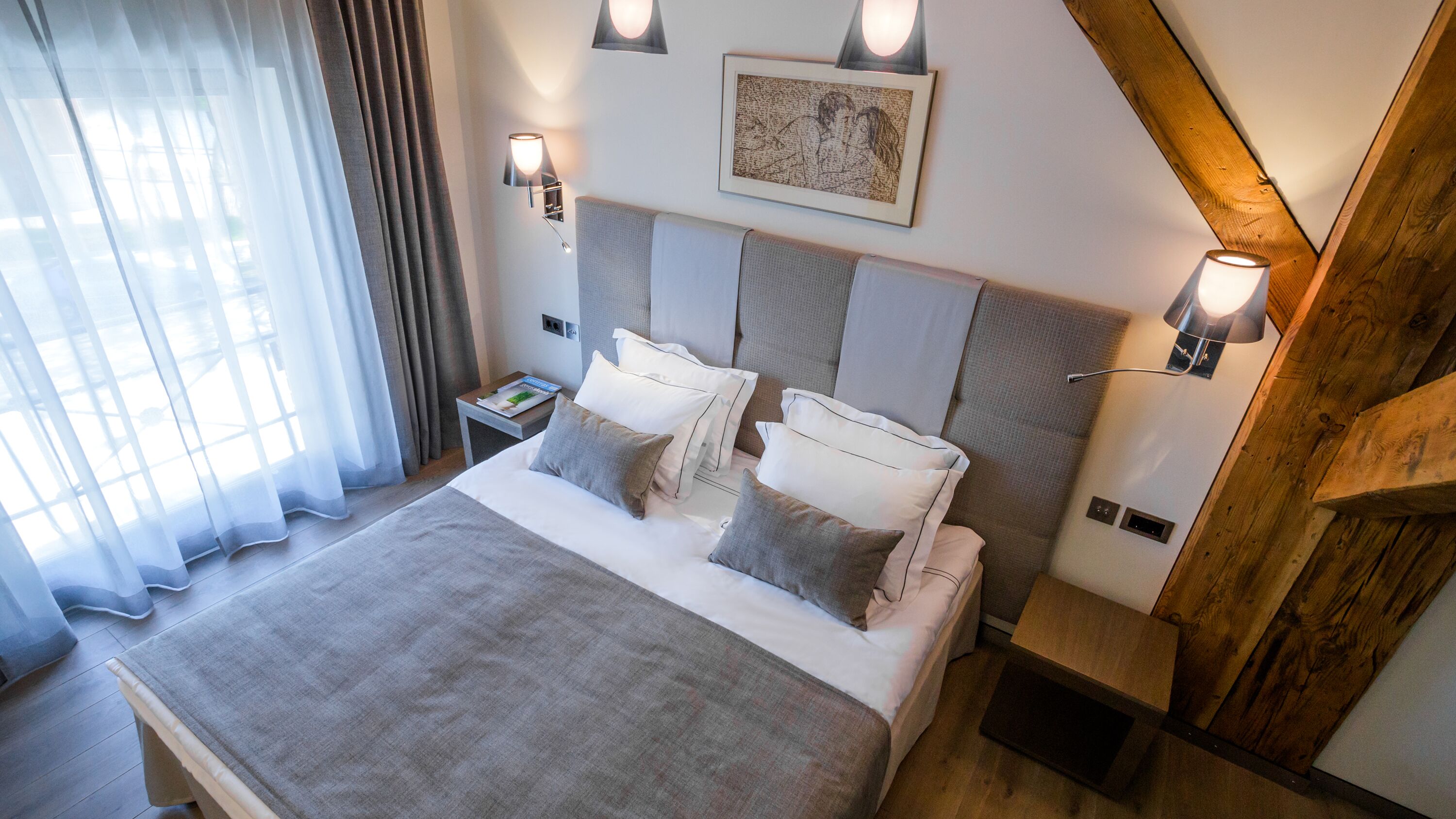 A modern hotel room with a large bed, a nightstand, a wall-mounted lamp, and a framed artwork above the bed.