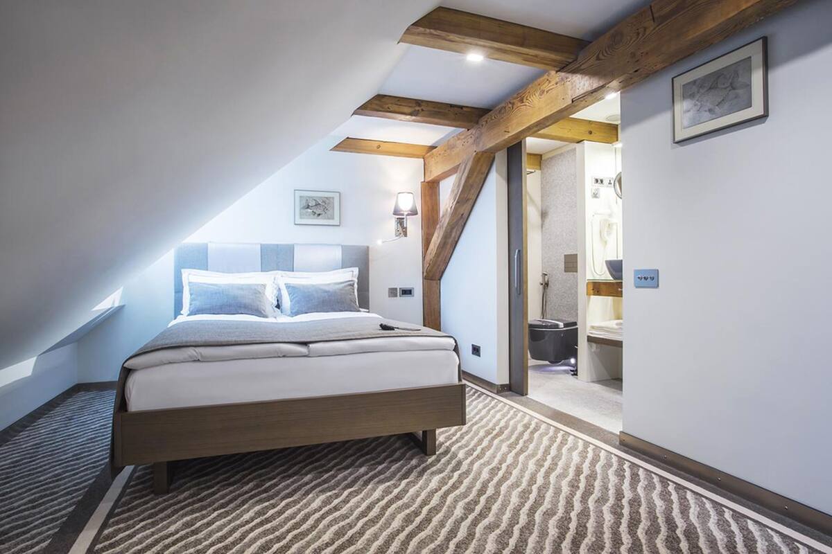 A bedroom with a bed, a wooden ceiling beam, a wall-mounted light, and a door leading to a bathroom.