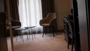 Quarto de hotel com uma janela grande, duas cadeiras, uma mesinha e uma escrivaninha com um computador.