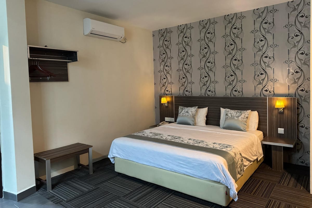 A hotel room with a bed, bedside lamps, a small table, an air conditioning unit, and a coat rack.