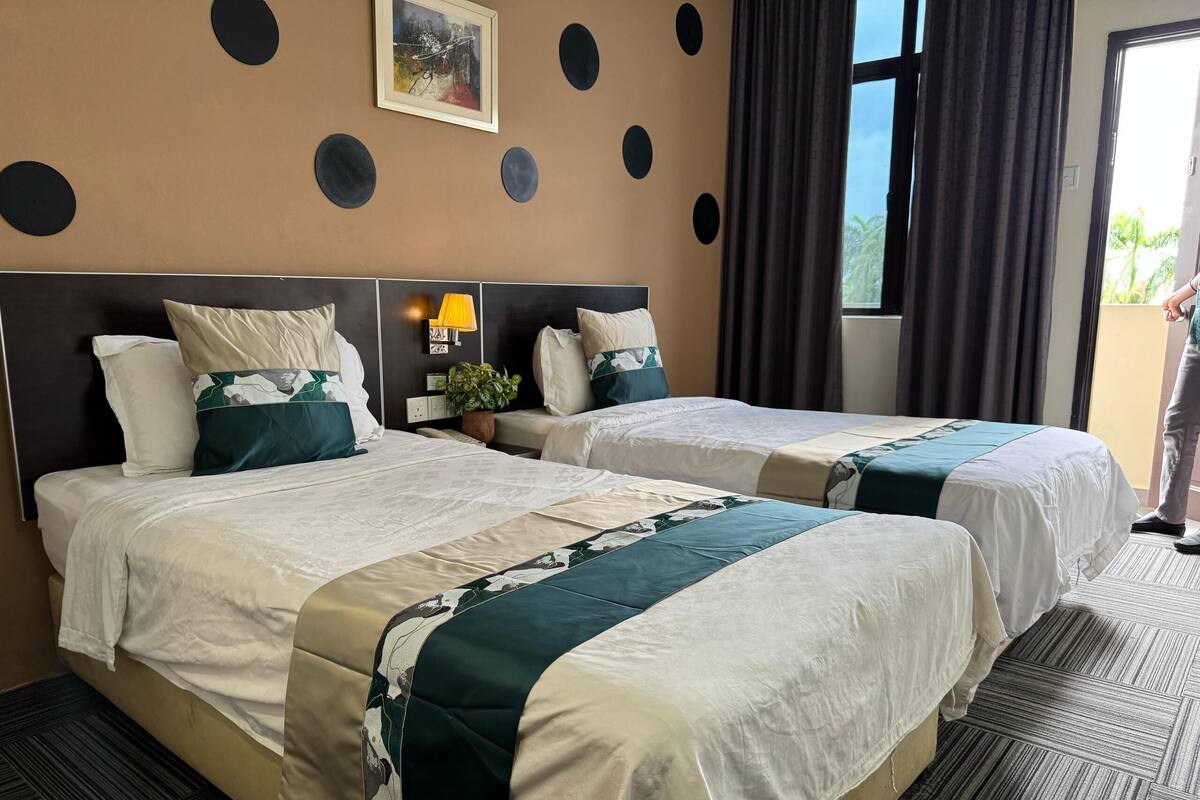 A hotel room with two beds, a wall-mounted lamp, a potted plant, and a window with curtains.