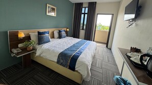 Habitación de hotel con una cama grande, una mesita de noche con lámpara, una planta pequeña en maceta, un escritorio con computadora y un cesto de basura.