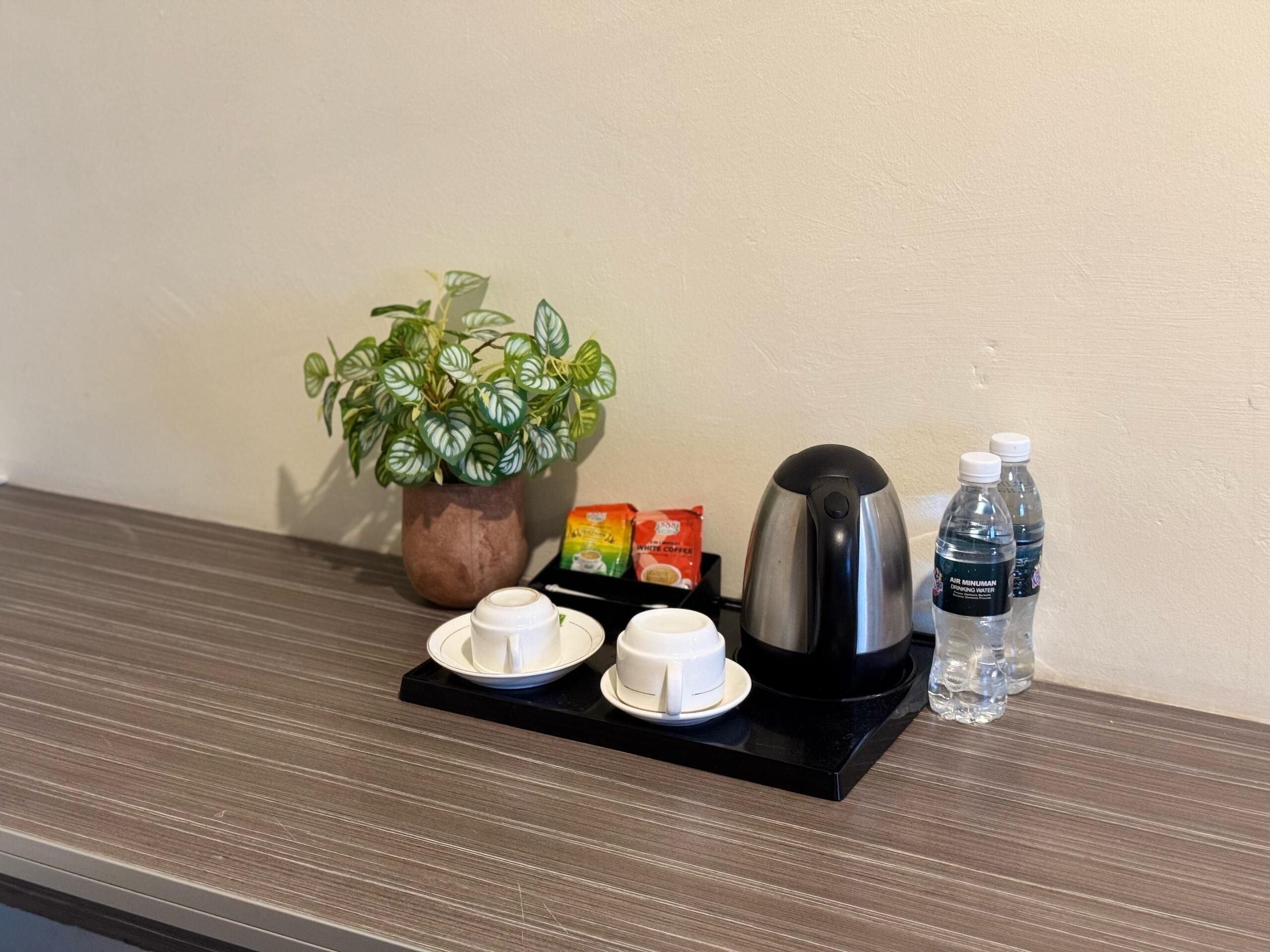A coffee station with a coffee maker, two cups, a packet of white coffee, a water bottle, and a potted plant.