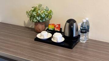 A coffee station with a coffee maker, two cups, a packet of white coffee, a water bottle, and a potted plant.