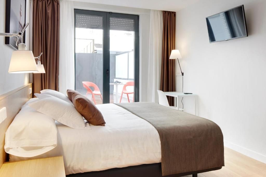 Habitación de hotel con cama, dos lámparas de noche, televisión montada en la pared y vistas al balcón exterior.