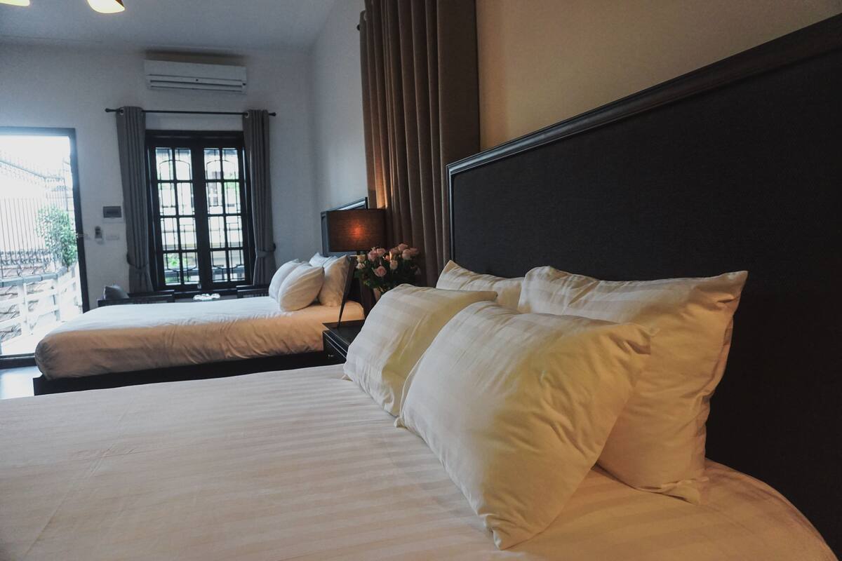 Ein Hotelzimmer mit zwei Betten, einem Nachttisch mit Lampe und Blick durch ein Fenster nach draußen.