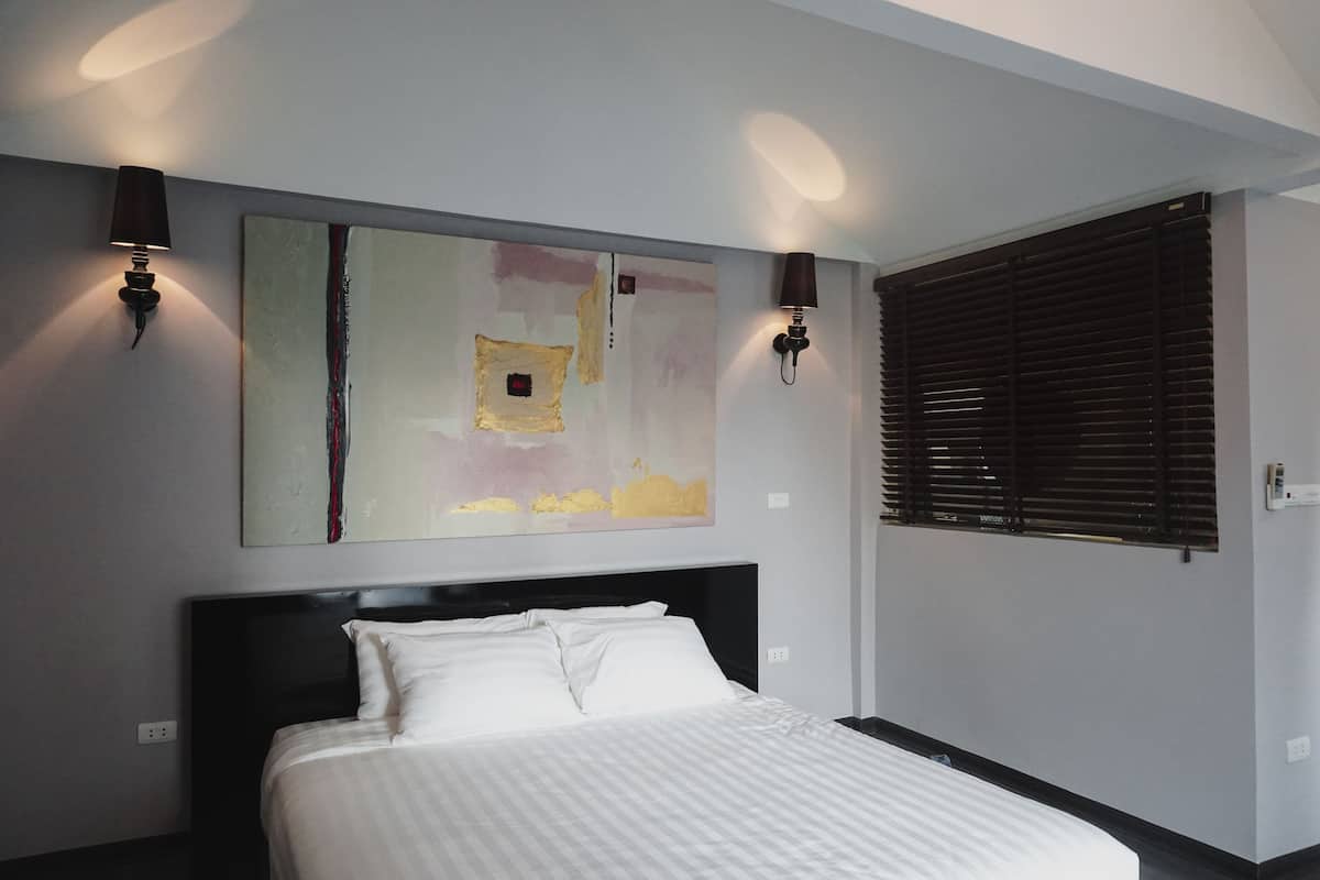 Ein Hotelzimmer mit einem großen Bett, zwei Wandlampen, einem Fenster mit Jalousien und einem großen, abstrakten Gemälde an der Wand.