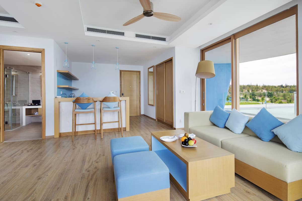 Una sala de estar moderna con un sofá, cojines azules, una mesa de centro con fruta y vistas al exterior.