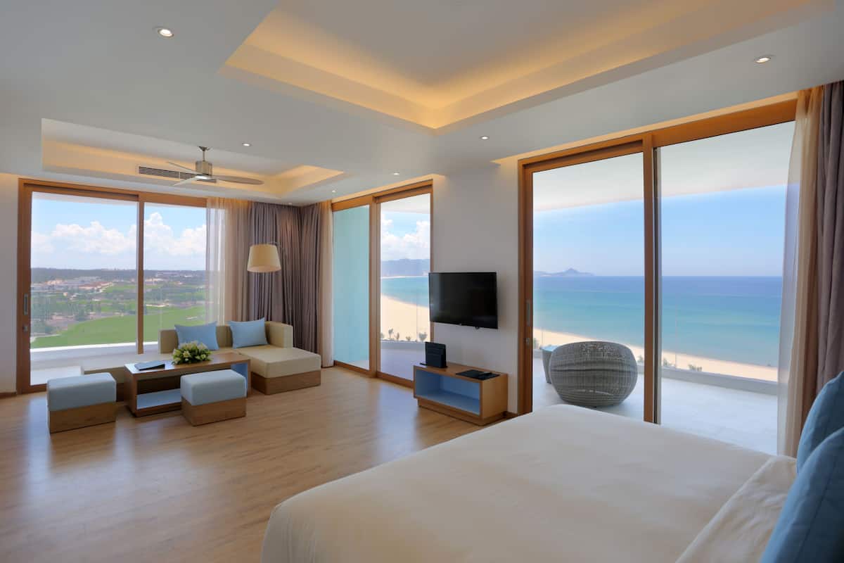 Una habitación de hotel moderna con una cama grande, un sofá, una mesita de café y una televisión. La habitación tiene ventanas amplias con vista a la playa y al océano.
