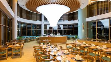 Een ruime eetruimte met lange houten tafels en groene stoelen met kussens, een bar in het midden en grote ramen voor natuurlijk licht.