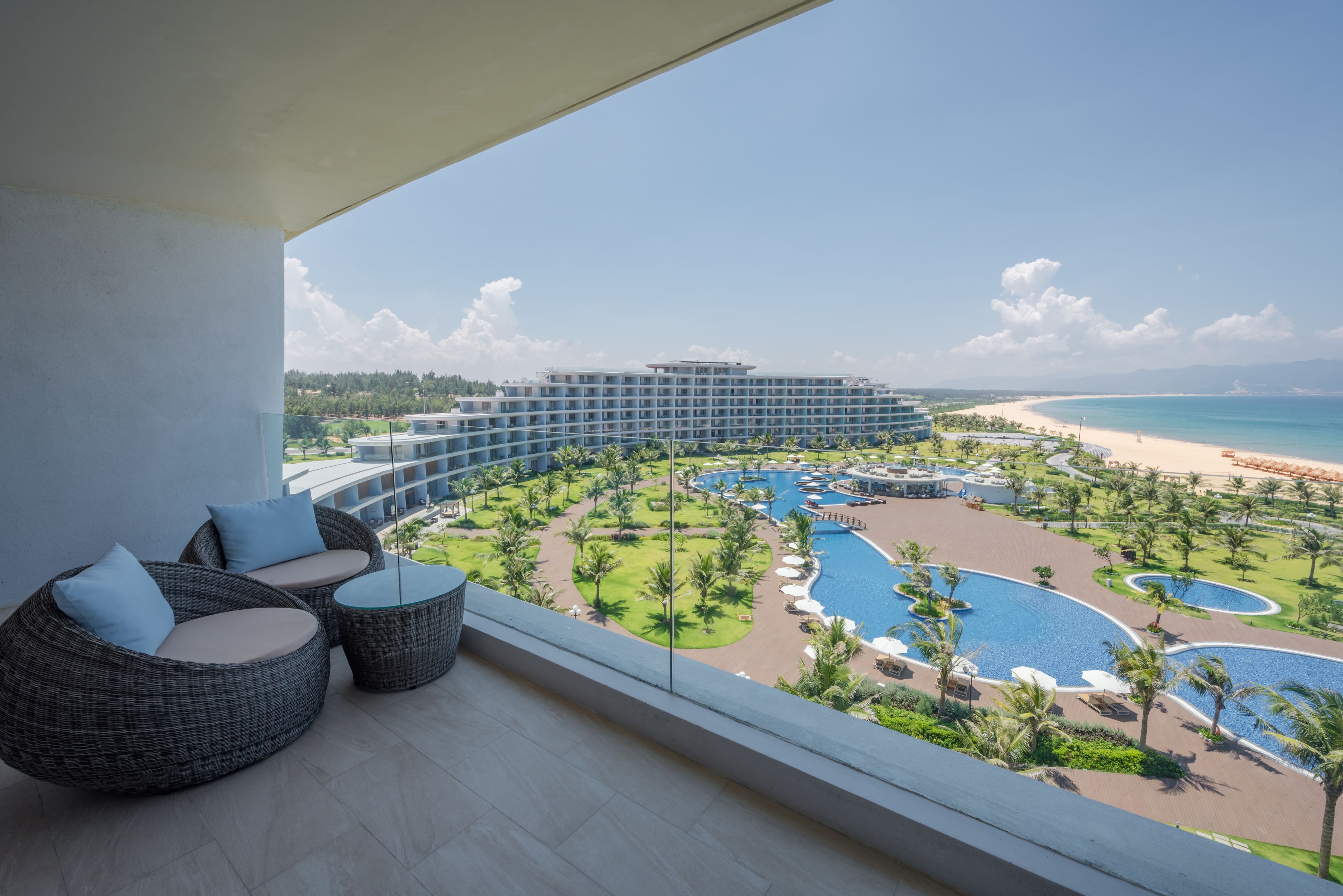 Un balcón con mobiliario de mimbre con vistas a una piscina de un resort, una playa y un edificio de hotel.