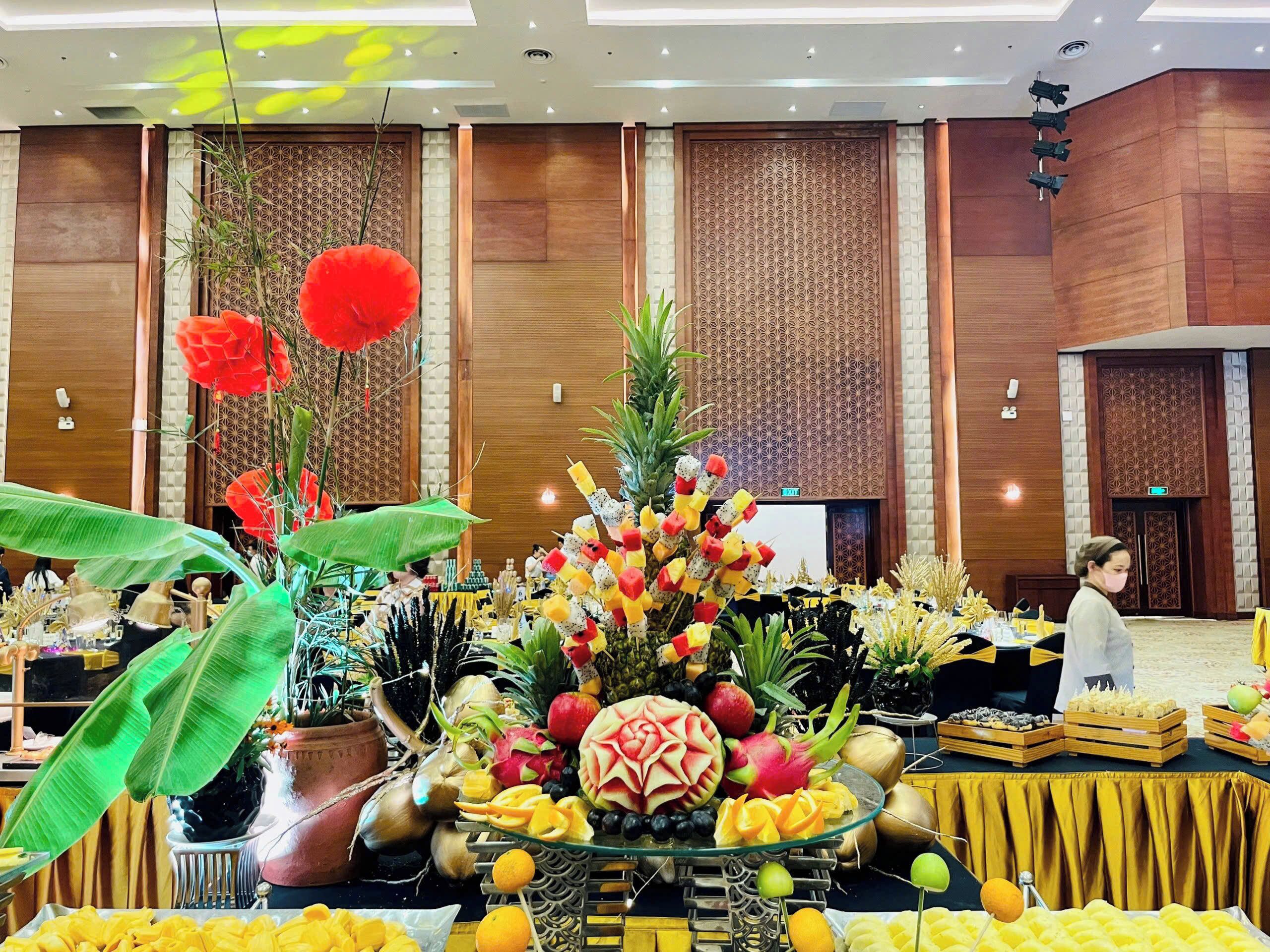 A buffet setup with various fruits and vegetables, a potted plant, and a decorative wall with a geometric pattern.
