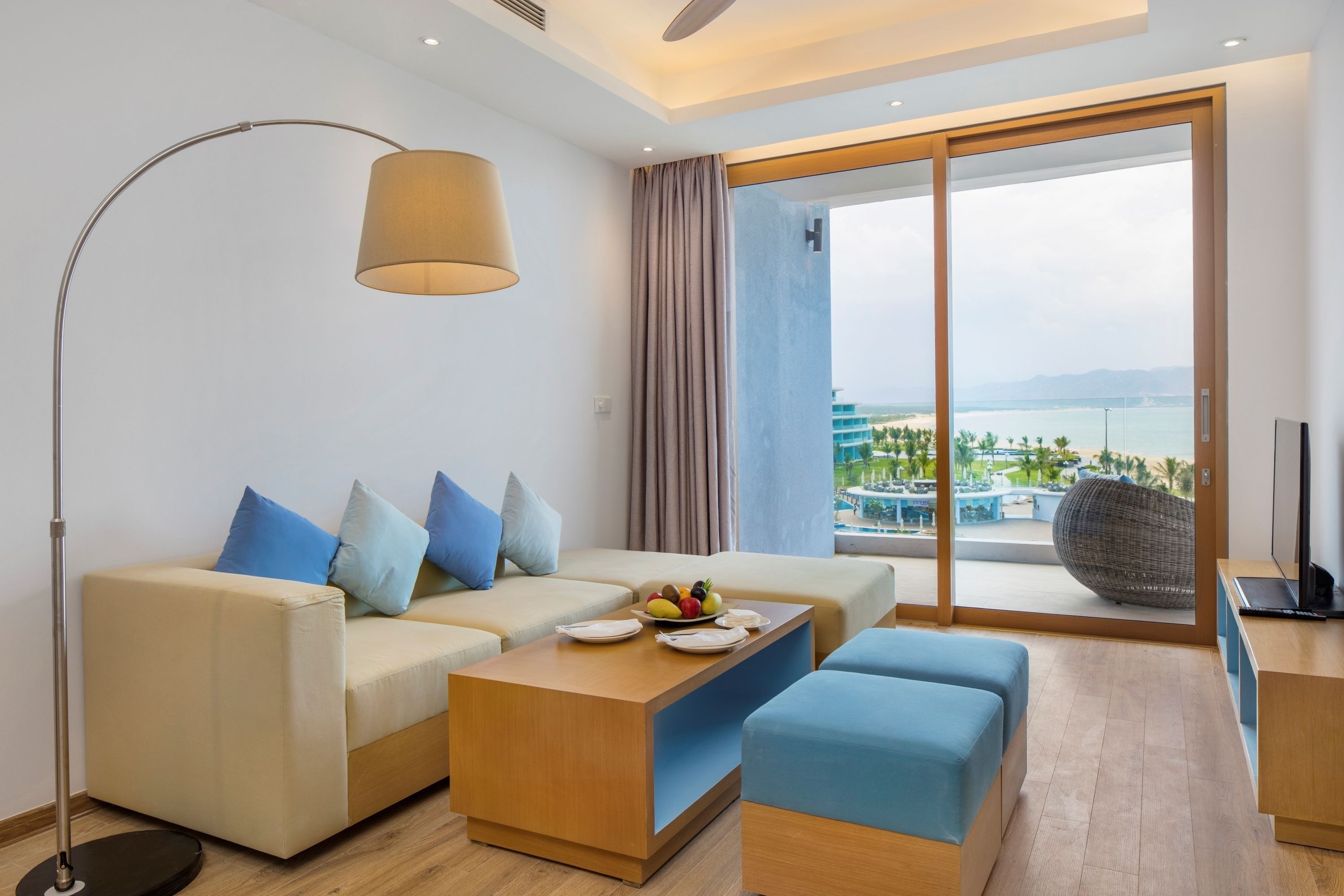 Une chambre d’hôtel moderne avec un canapé, une table basse sur laquelle sont disposés des fruits, une télévision et une grande fenêtre donnant sur une piscine et des montagnes.