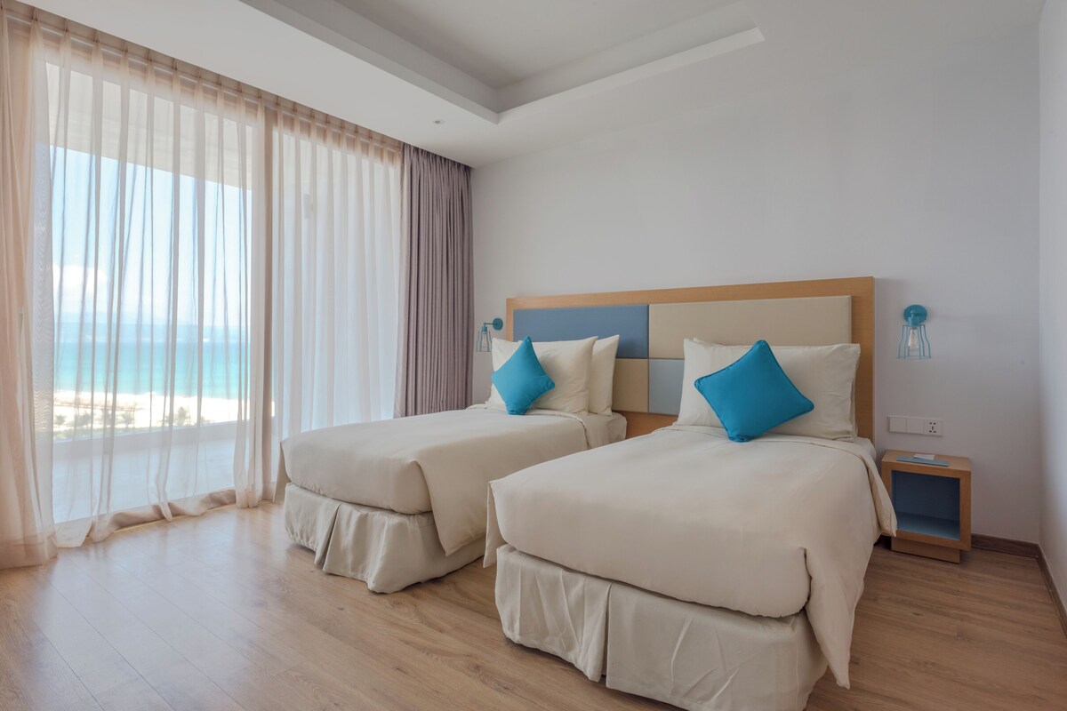 A hotel room with two beds, a wooden floor, a nightstand, and a view of the sea.