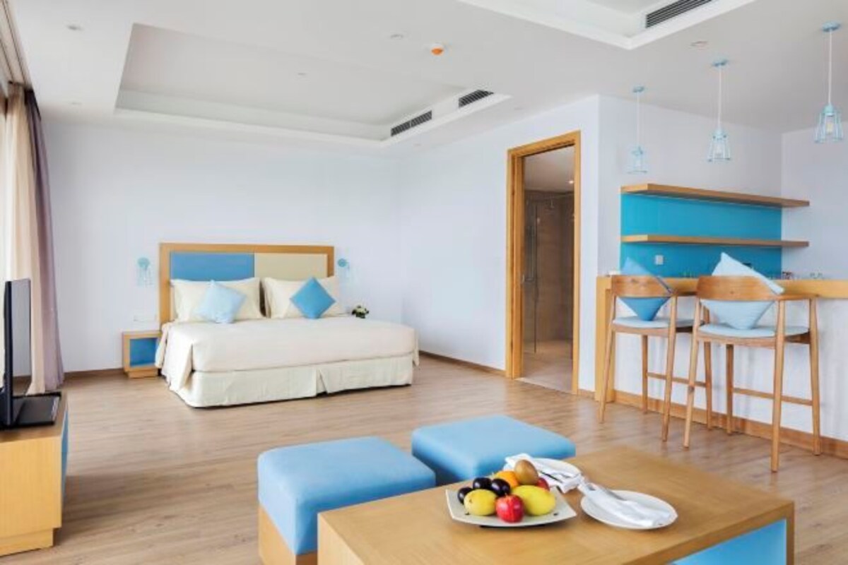 A modern hotel room with a bed, a dining area with bar stools, and a small table with fruit.