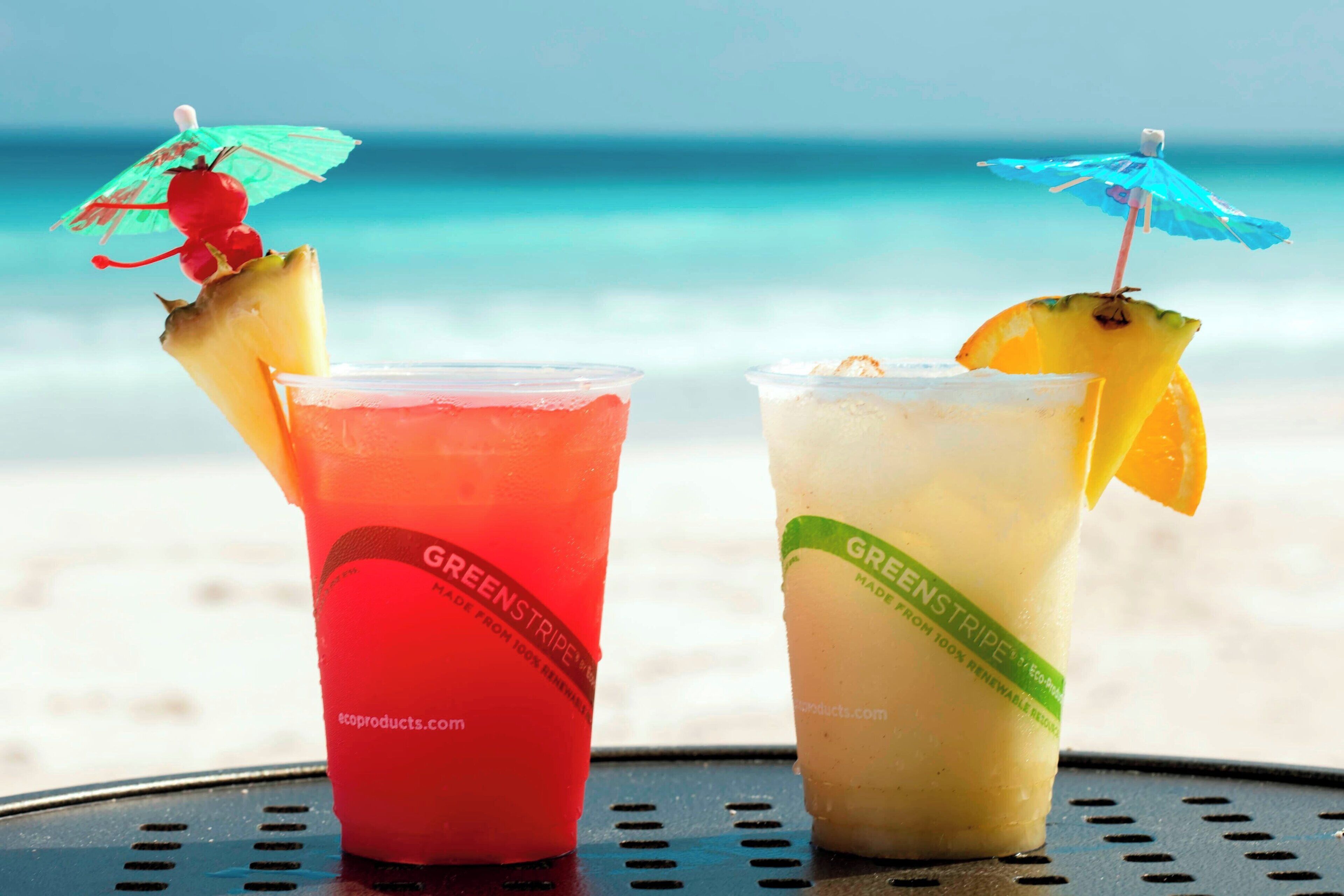 Two drinks with fruit garnishes on a table by the beach.