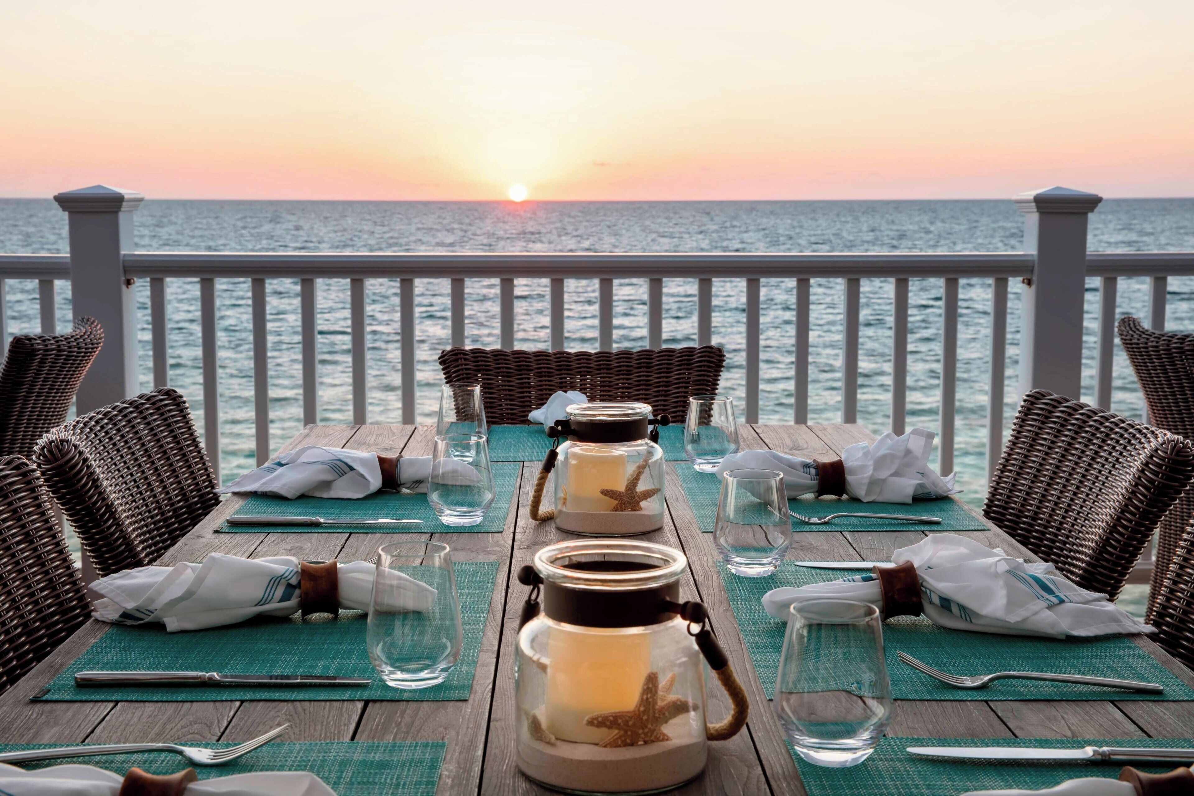 Una mesa de comedor dispuesta para dos personas, con sillas de mimbre, un candelabro de cristal y vistas al océano al atardecer.