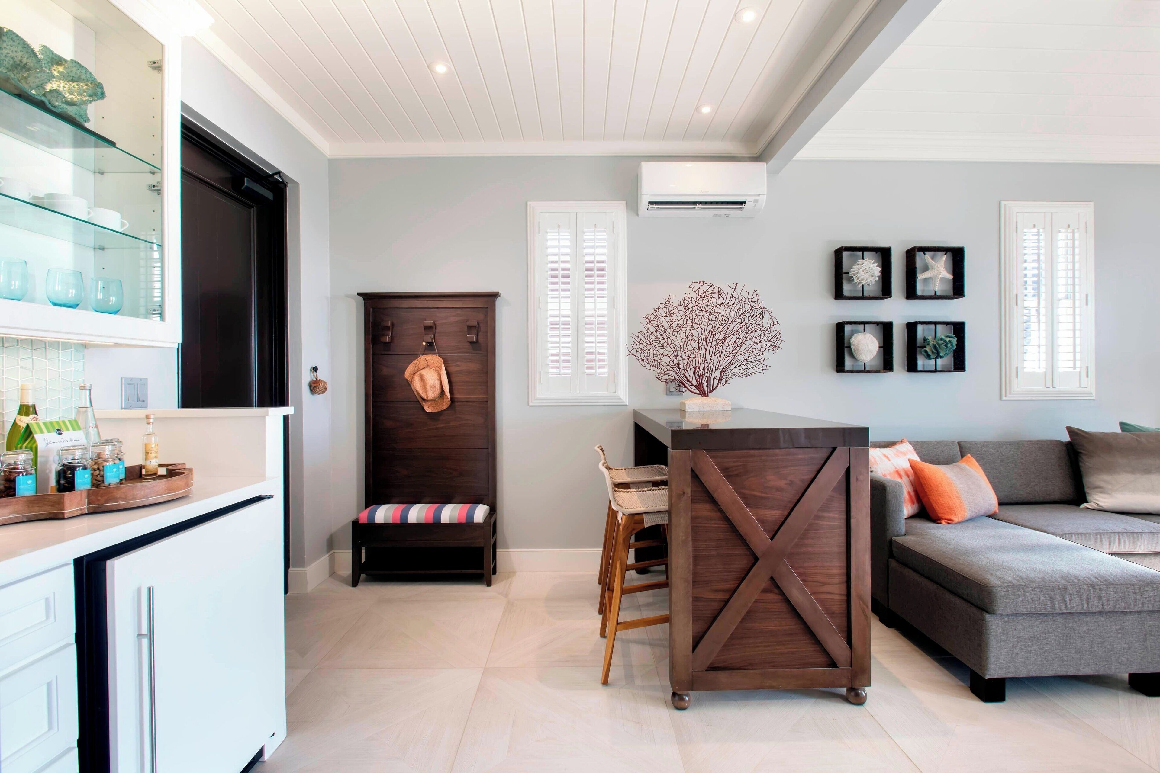 A modern living room with a grey sectional sofa, a wooden dining table, and a kitchen area with white cabinets and a glass display shelf.