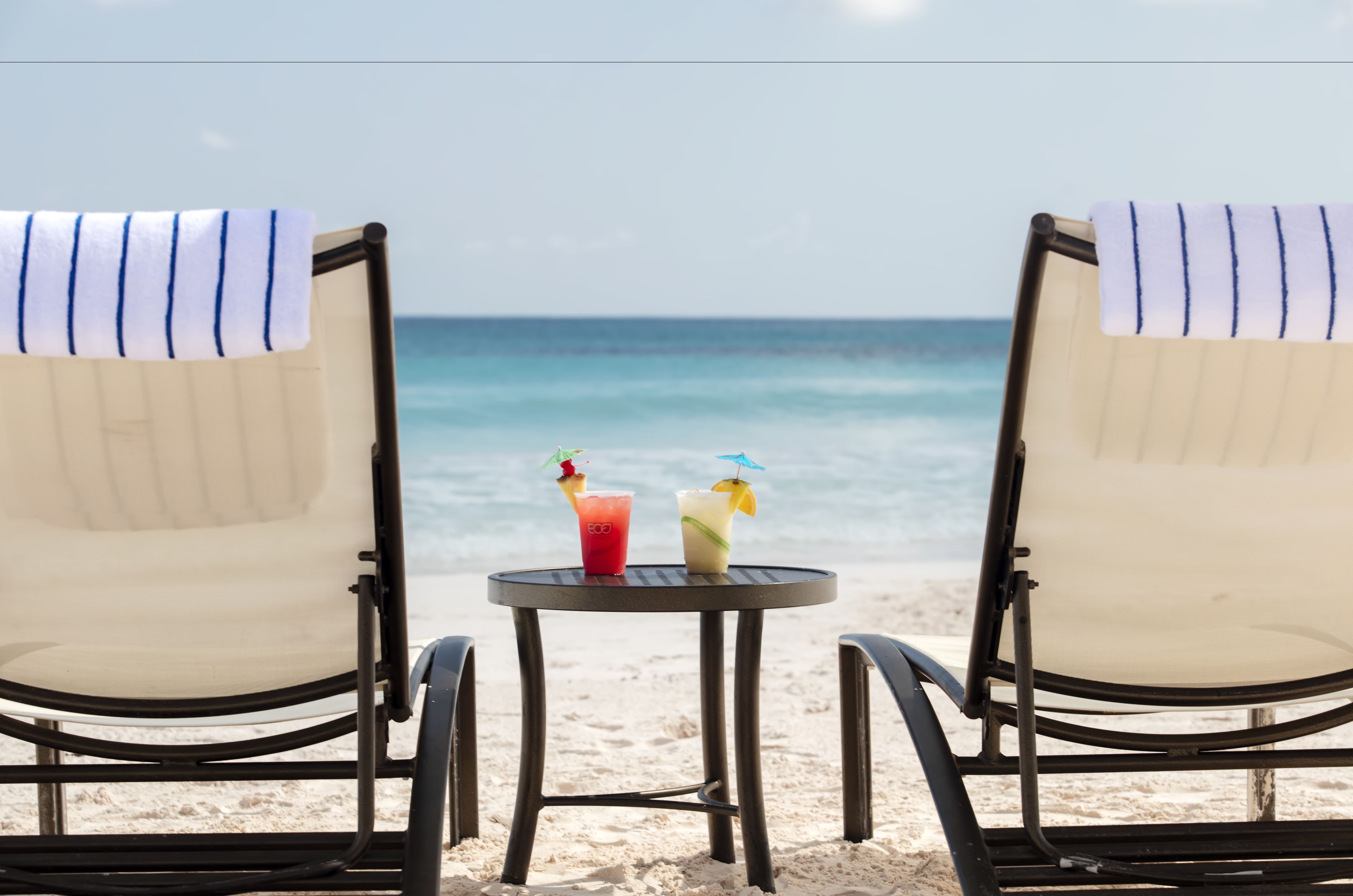 Deux chaises de plage avec des serviettes à rayures, une petite table ronde avec deux boissons, et une vue sur l’océan.