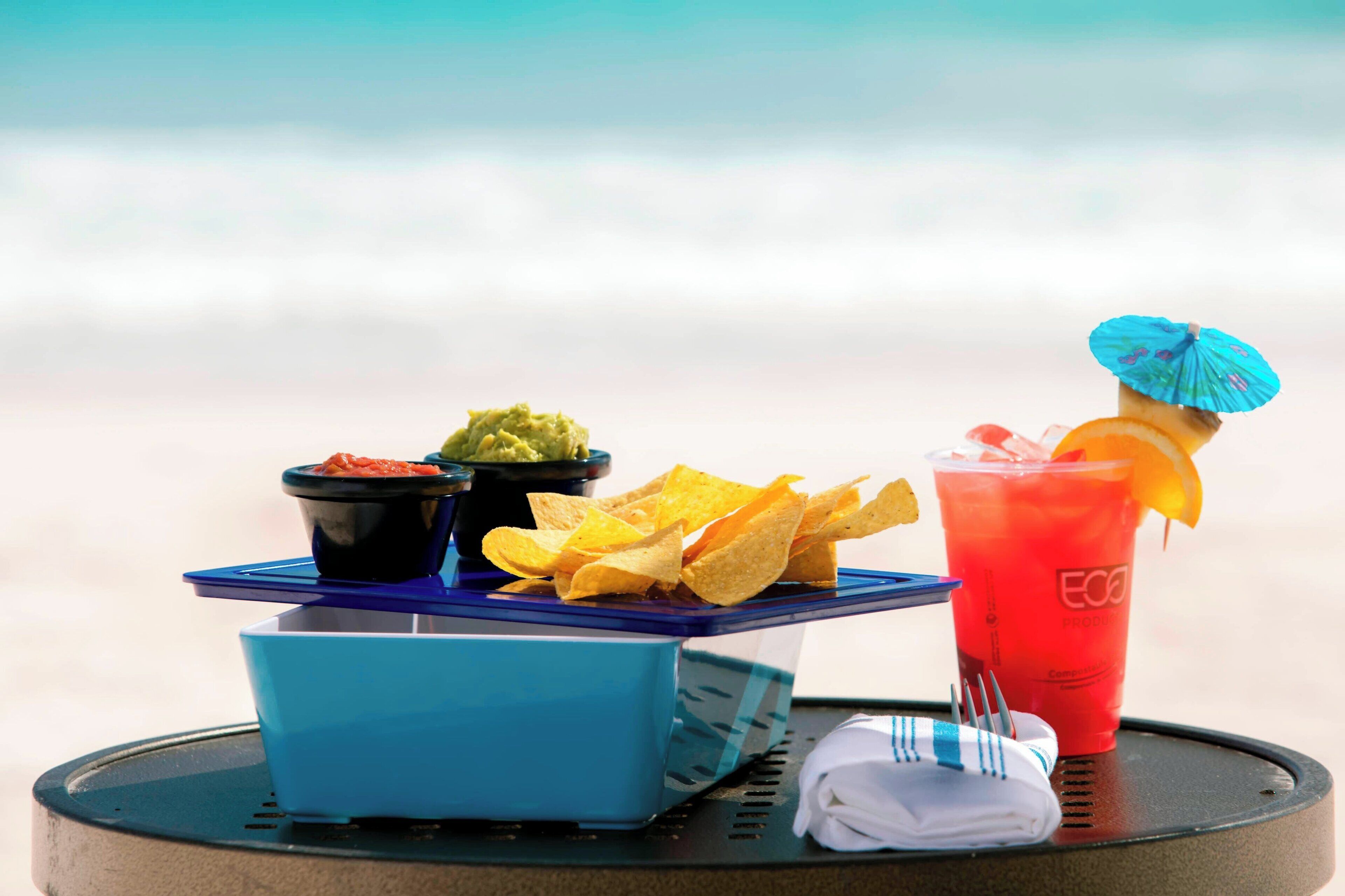 A table with a blue tray holding nachos, a red drink with an orange garnish, and two small bowls of dips.