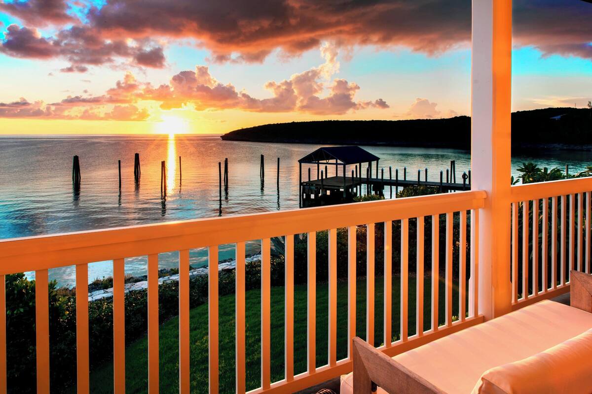 A balcony with a view of the sunset over the water, a wooden deck, and a covered area.