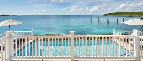 A pool with a white railing and two umbrellas, overlooking a clear blue sea with a distant island.