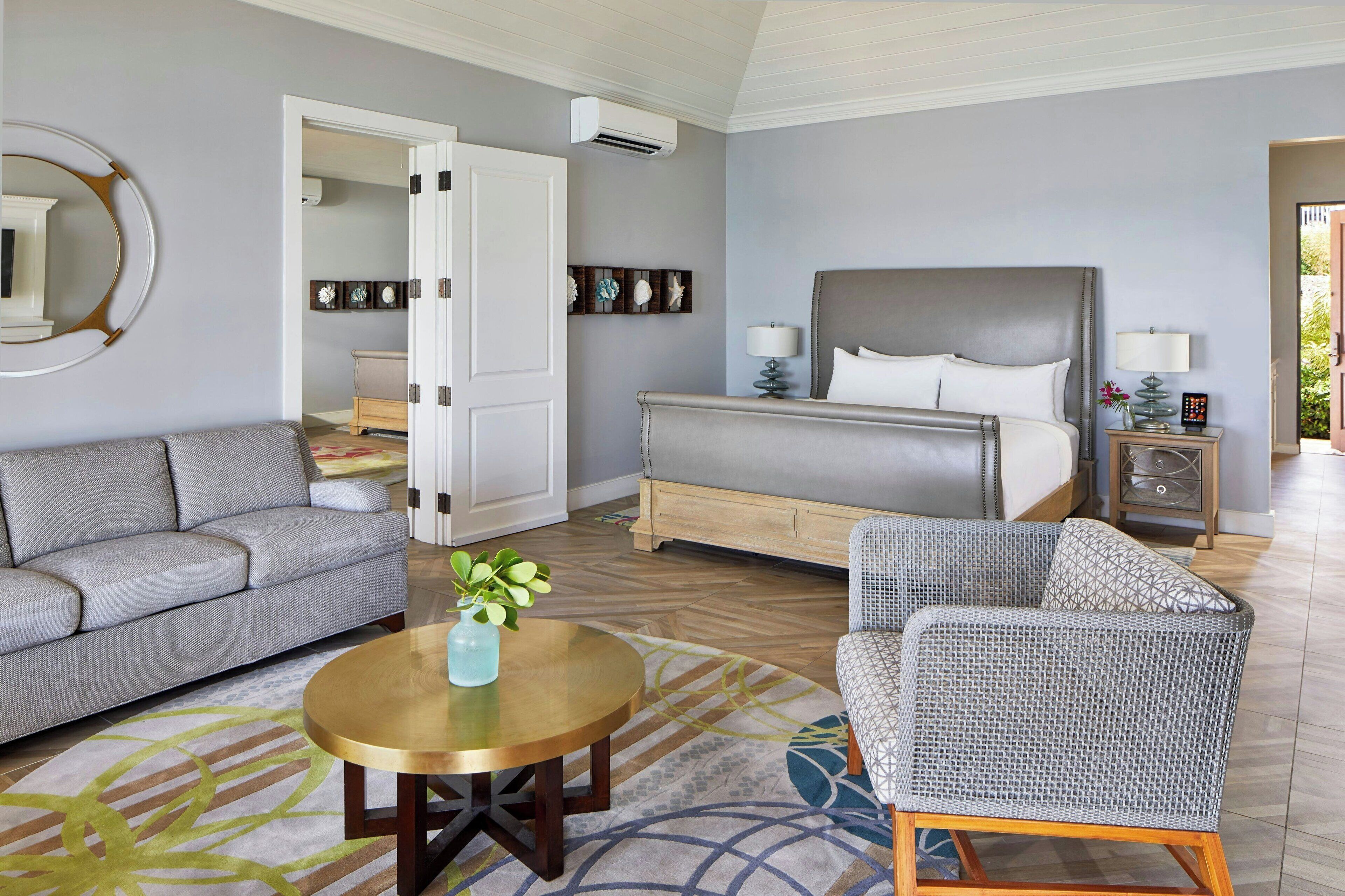 Quarto de hotel com cama, sofá, poltrona, mesa redonda e uma pequena planta.