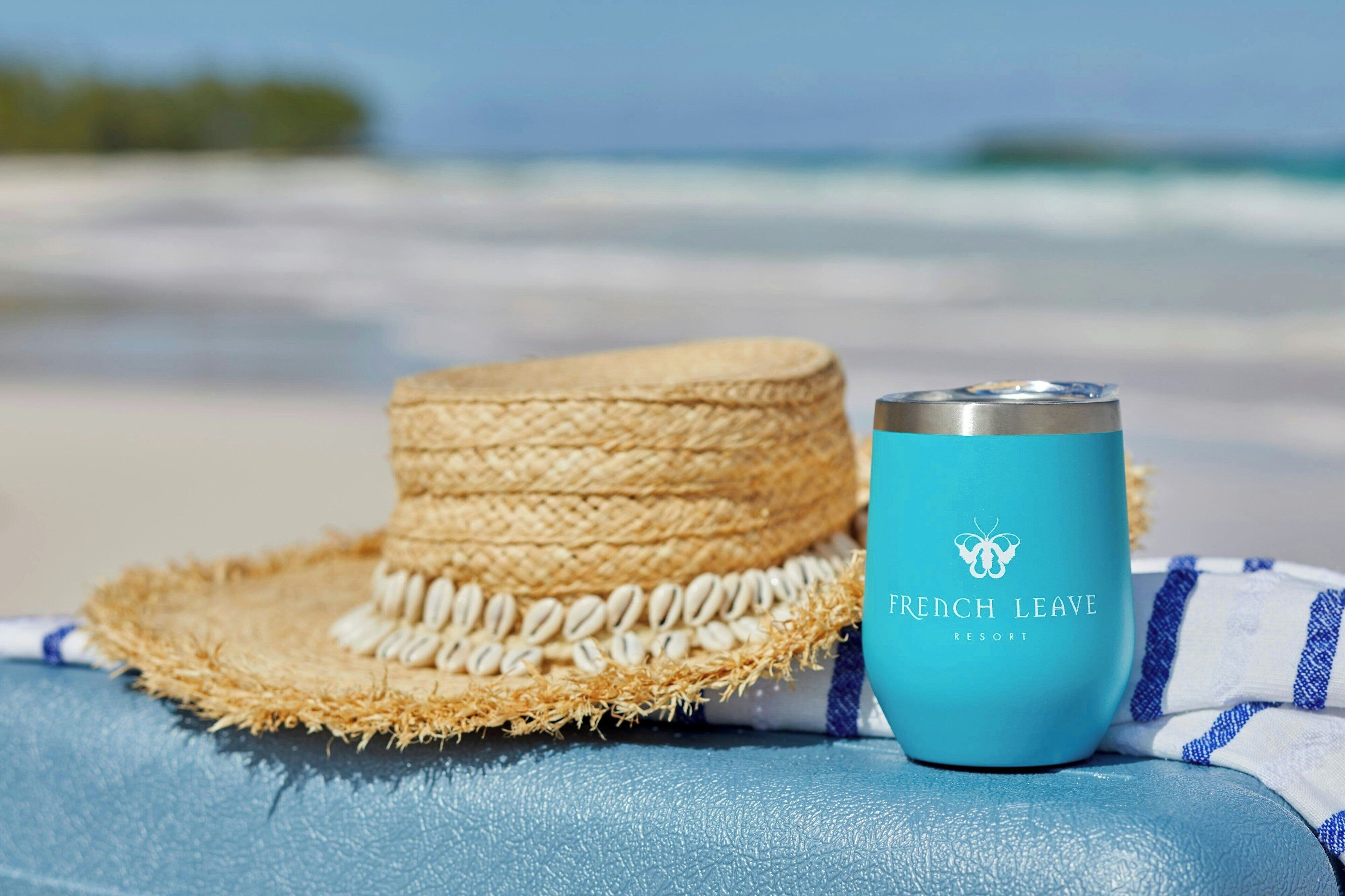 Un chapeau de paille et un verre bleu sur lequel est inscrit « French Leave Resort », le tout sur une plage.