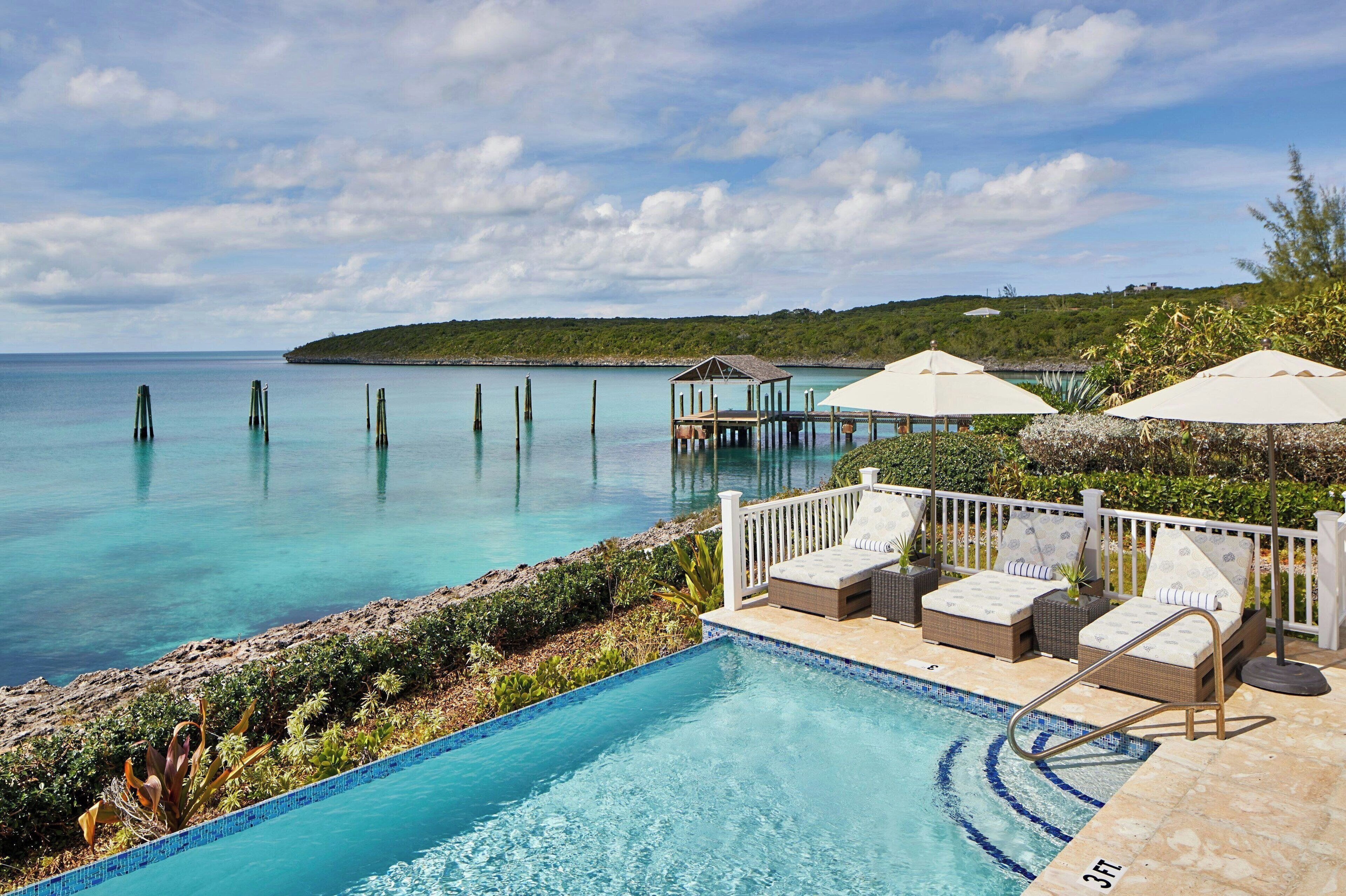 A poolside area with lounge chairs and umbrellas, overlooking a clear blue sea with wooden posts.