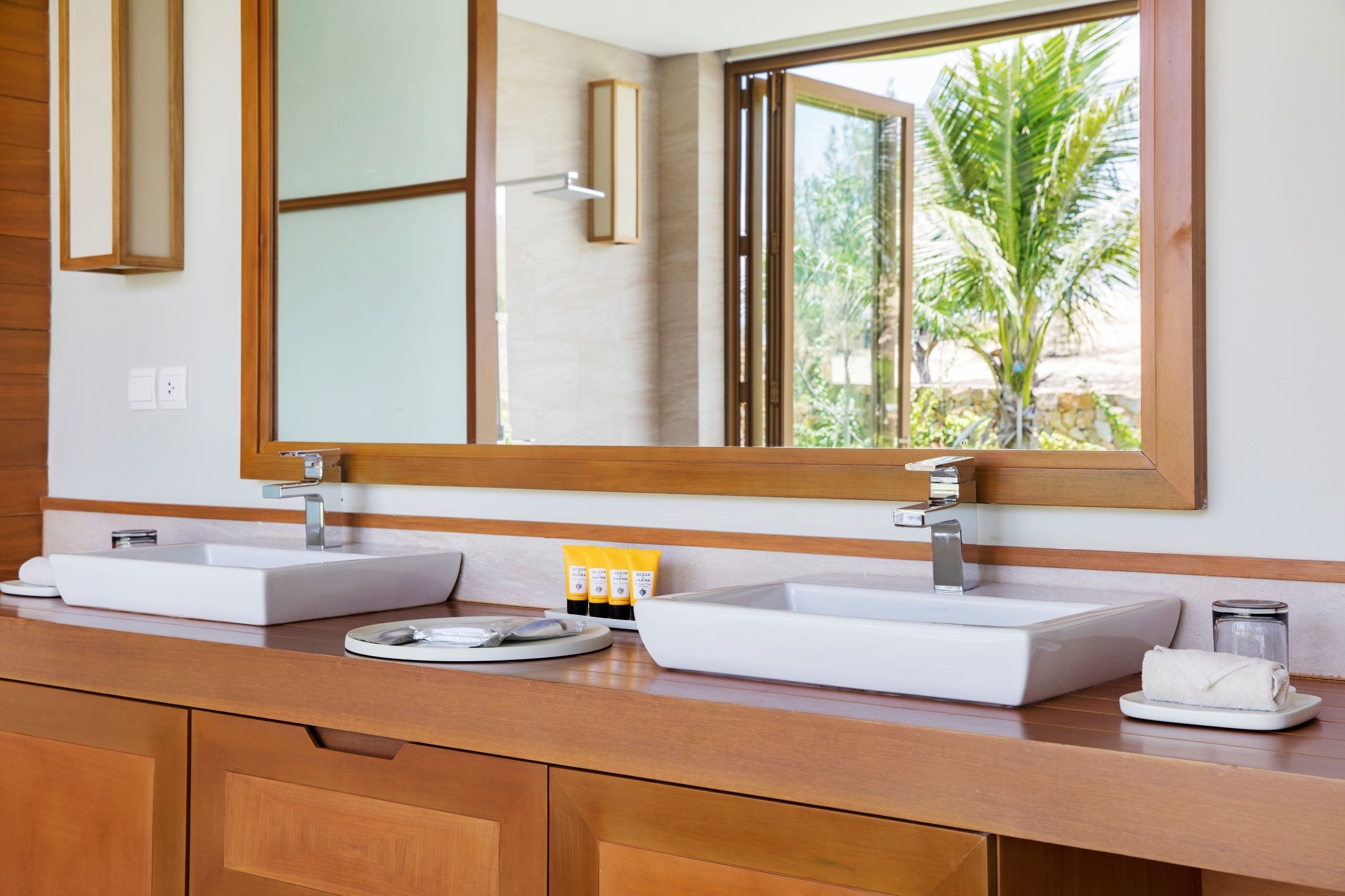 A bathroom with two sinks, wooden cabinets, a large mirror, and a window with a view of palm trees.