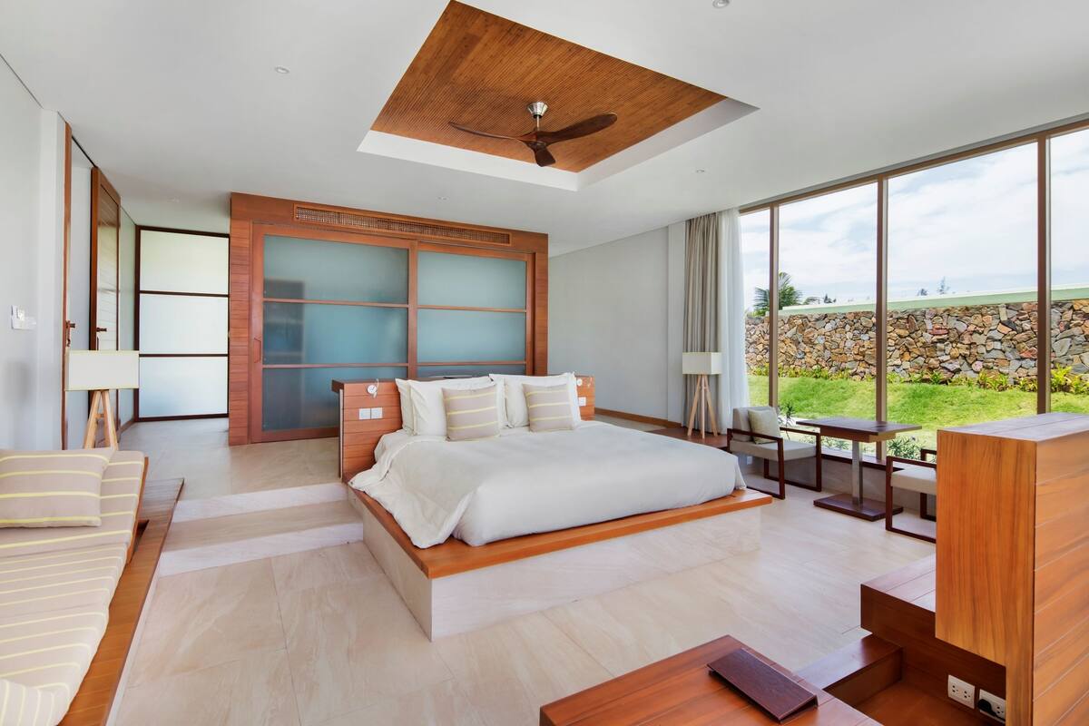 Une chambre moderne avec un grand lit, un ventilateur de plafond en bois et une vue sur un mur de pierre et la verdure environnante.