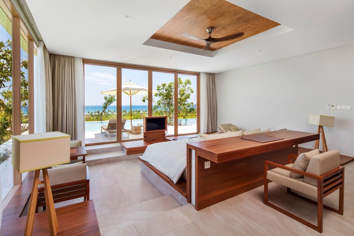Une chambre d’hôtel moderne avec un bureau en bois, un lit, un canapé et une télévision. Une grande fenêtre offre une vue sur l’océan et une piscine.