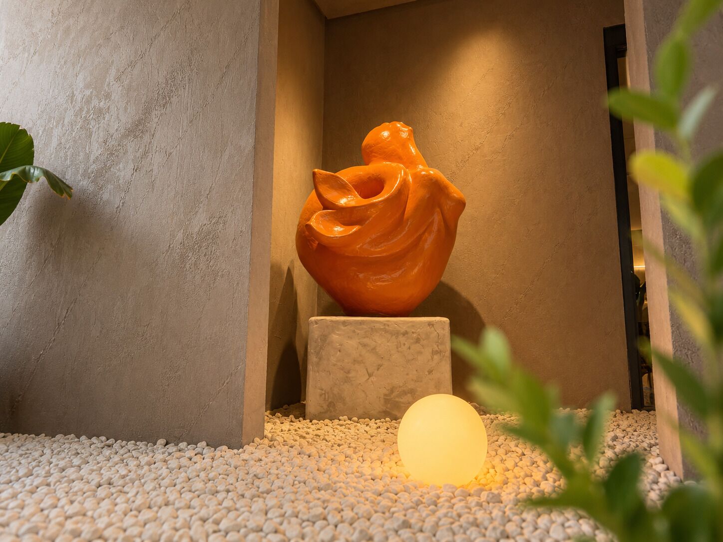 Un intérieur moderne agrémenté d’un vase orange aux formes sculpturales, posé sur un piédestal, d’un luminaire sphérique diffusant une lumière douce et d’un sol recouvert de petits galets.