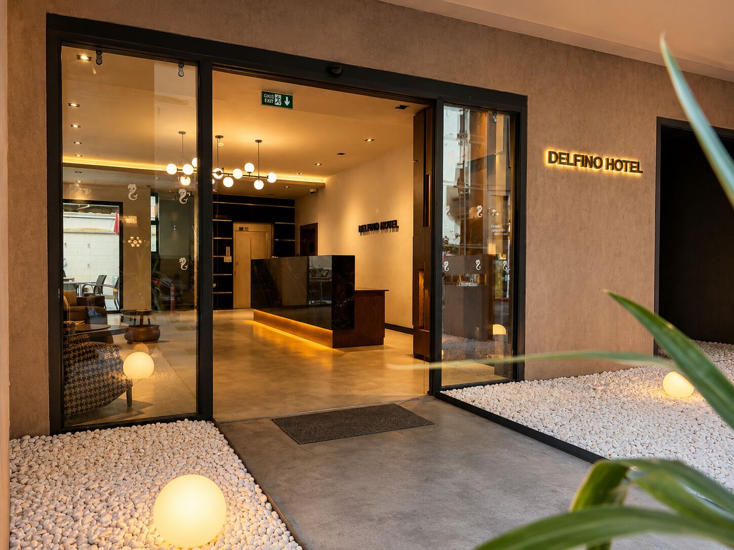 A hotel entrance with a reception desk, a sign indicating an exit, and a modern interior with a sofa and decorative lighting.