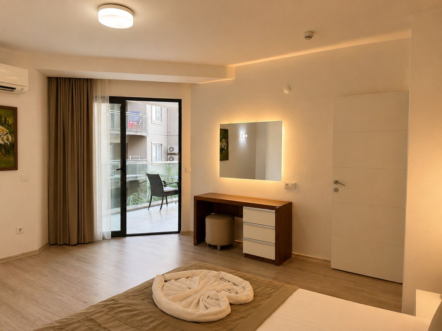 Een moderne hotelkamer met een bed, nachtkastje, spiegel, een schuifdeur naar een balkon en uitzicht op de omgeving.