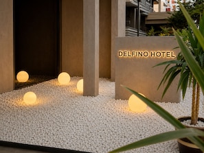 A hotel entrance with a sign reading 'DELFINO HOTEL,' surrounded by white pebbles and illuminated spherical lights.