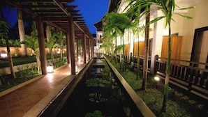 A nighttime view of a hotel corridor with a water feature, palm trees, and a covered walkway.