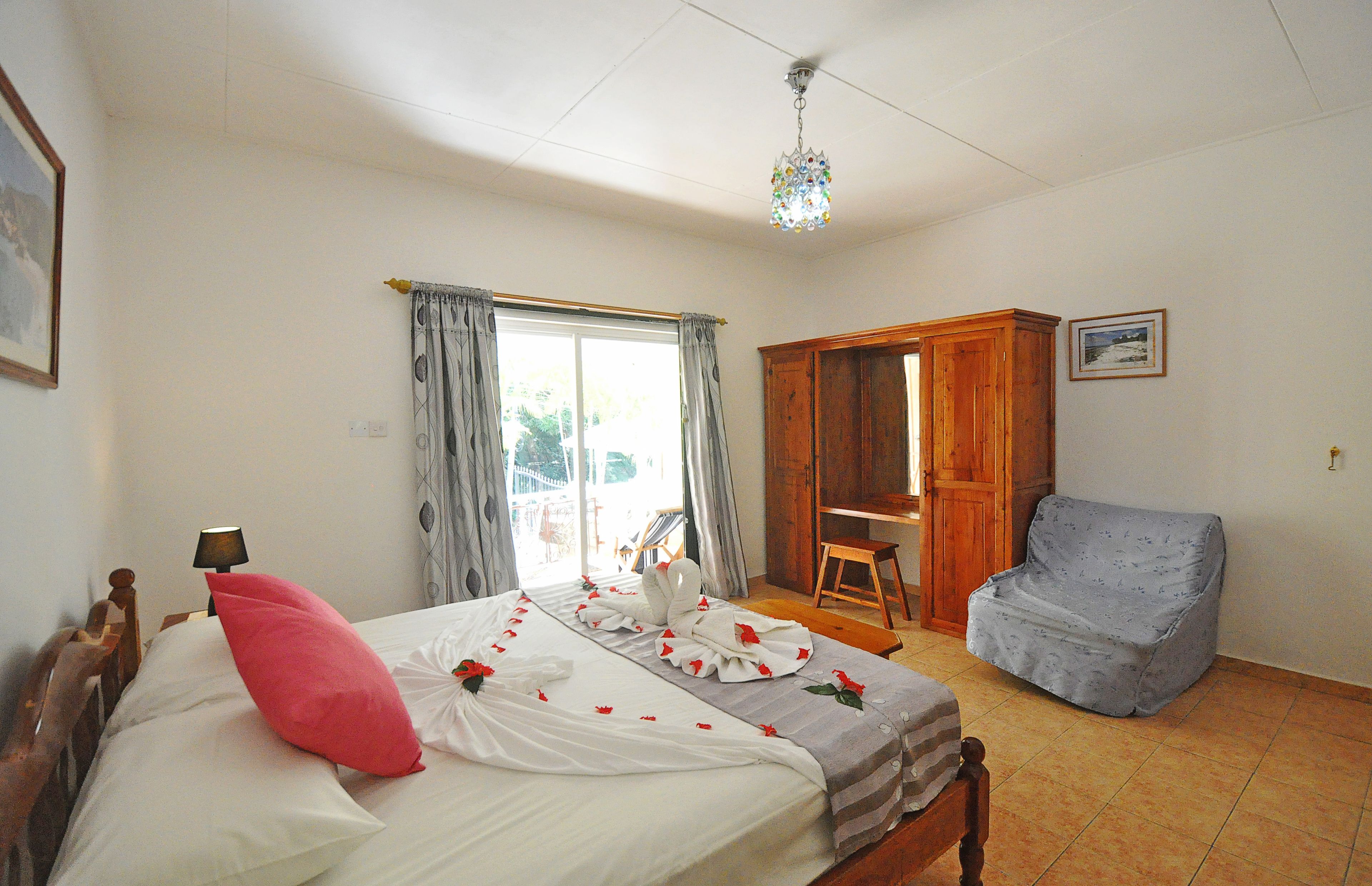 A bedroom with a bed, a wooden wardrobe, a covered chair, and a window with curtains.