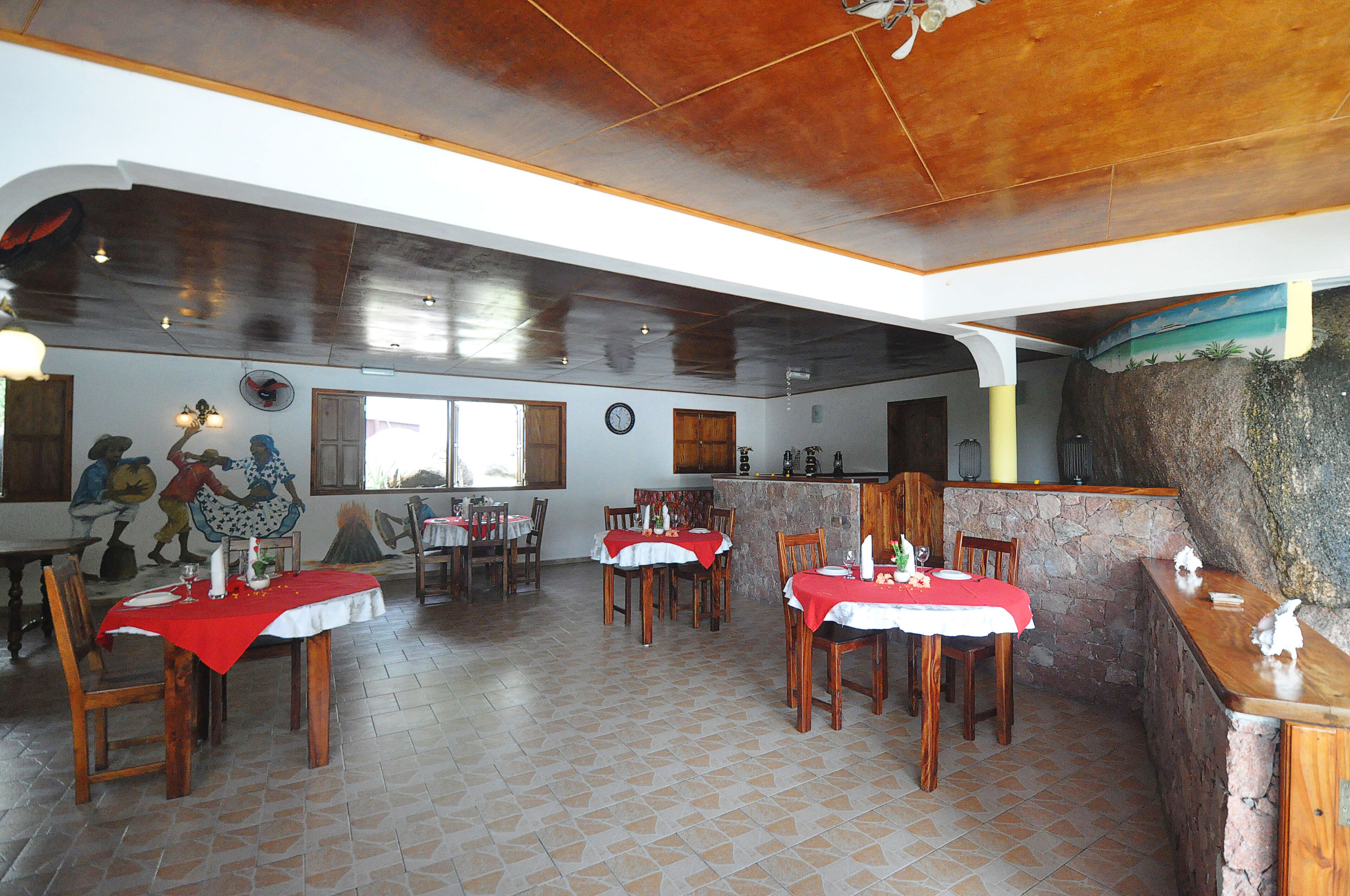 A restaurant with multiple tables, wooden chairs, and a stone wall.